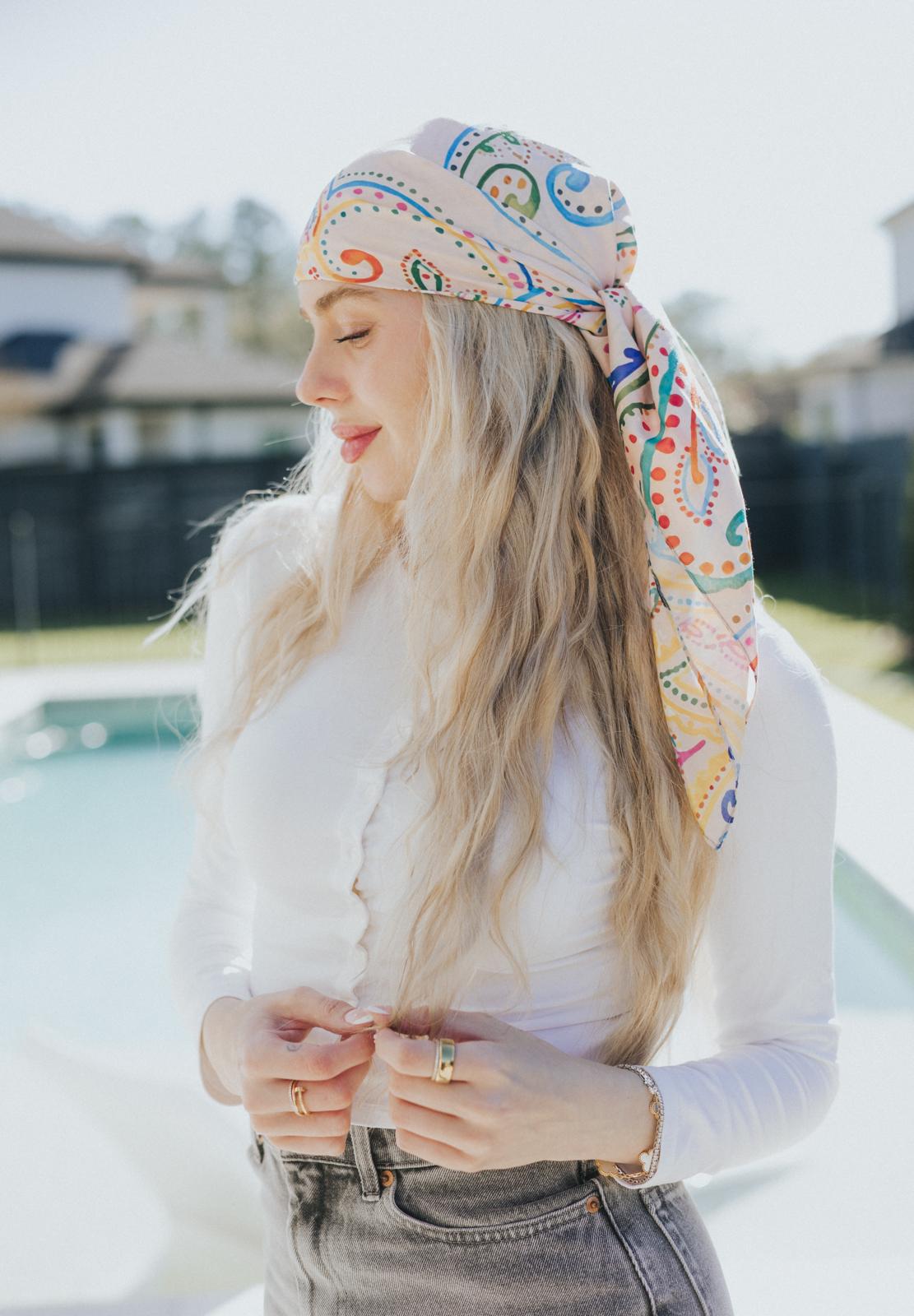 Woman with long wavy blonde hair wearing a silk headscarf in a playful paisley pattern of multicolored swirls and dots, tied at the back and styled with a white top and gray jeans in an outdoor setting by a pool.