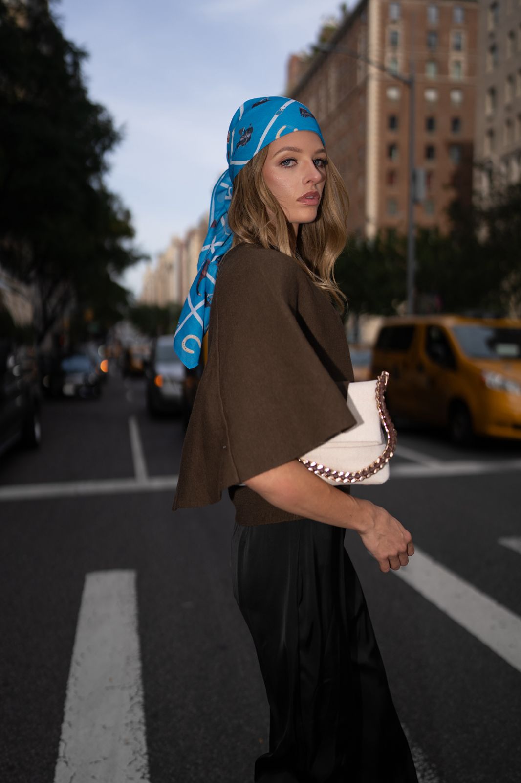 Model wearing TUK’s Blue Reign headscarf styled over brown top and black pants, city street fashion look with chic elegance.