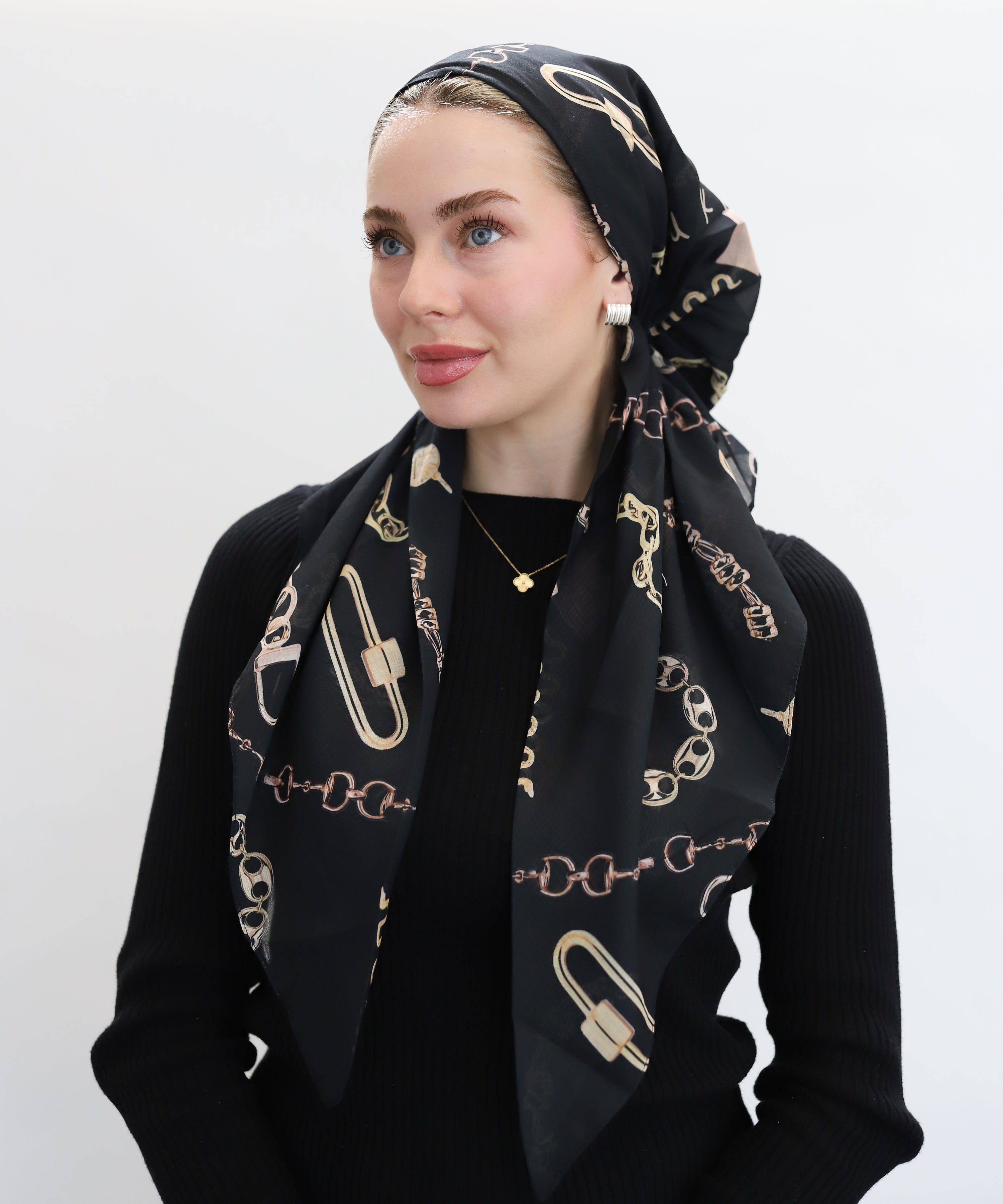 Woman wearing the Charmed Pre-tied Head Scarf by TUK features a bold black canvas adorned with intricate gold chain motifs, padlocks, charms, and ornamental links, arranged in a diagonal grid pattern for maximum visual impact. 