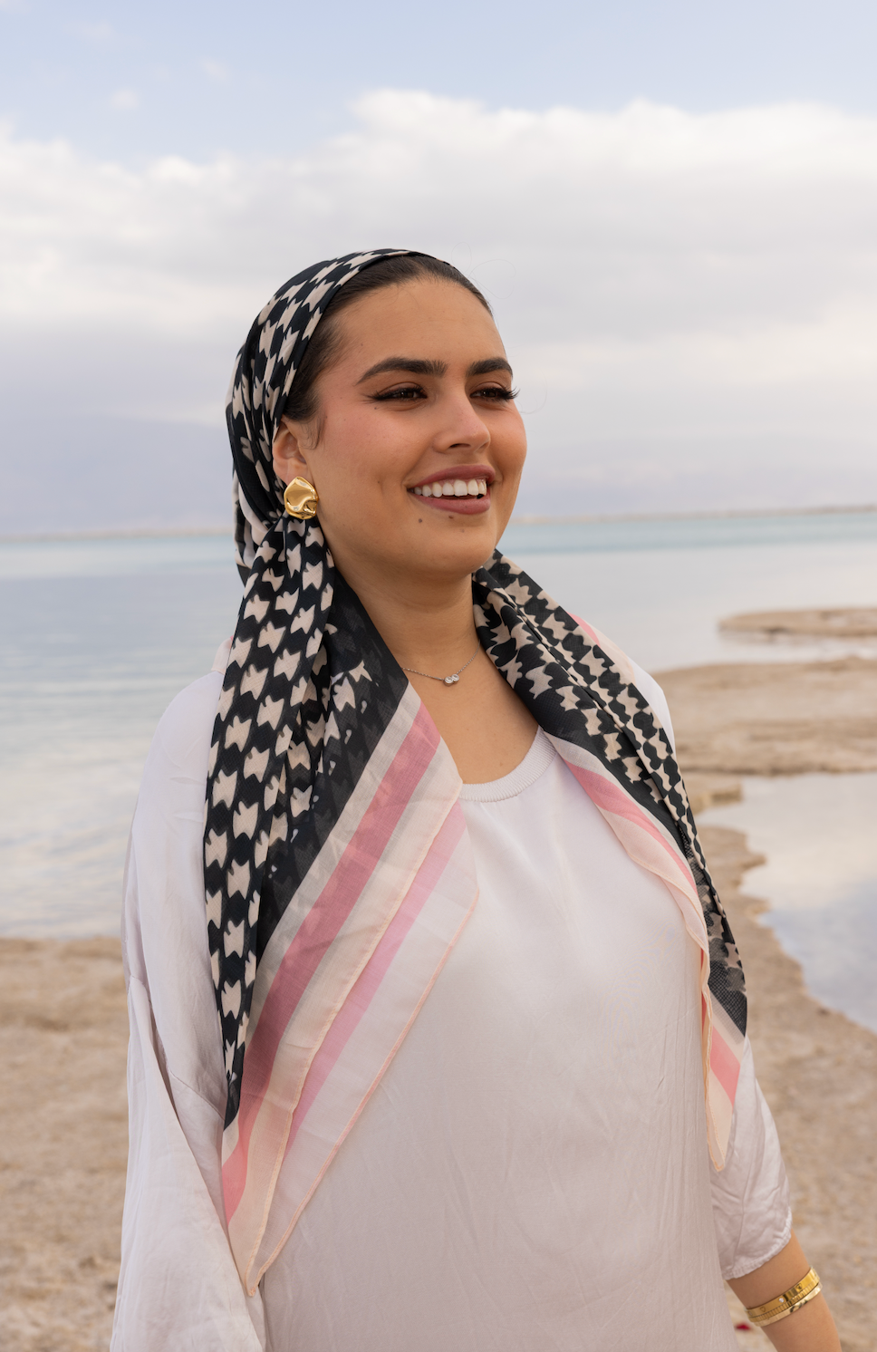Woman wearing black and cream houndstooth Checked Out headscarf with soft pink border, styled elegantly by the sea.