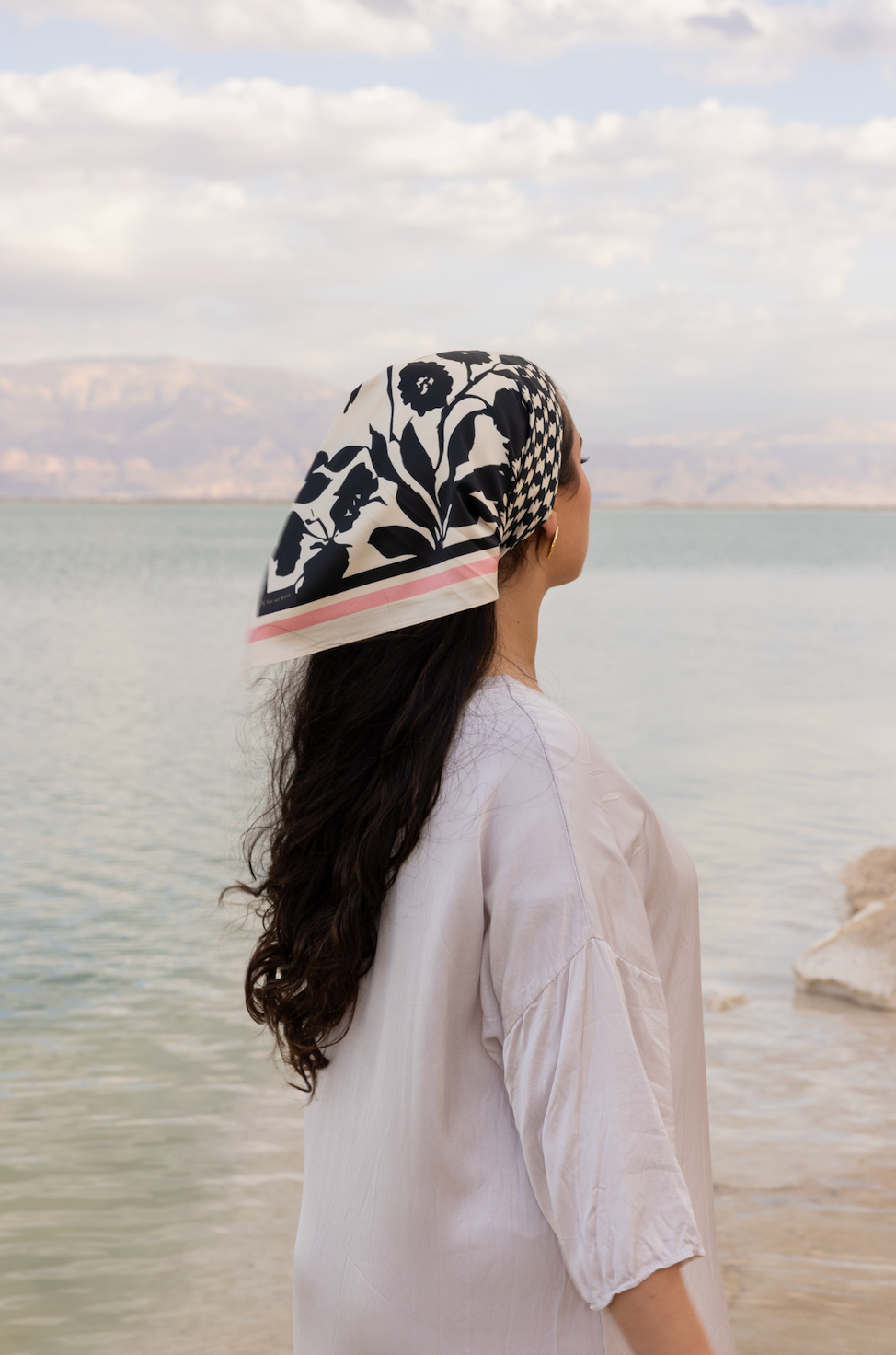 Model by a calm seaside wearing TUK Checked Out Silk Scarf, black floral print with pink edge, styled over flowing long hair.
