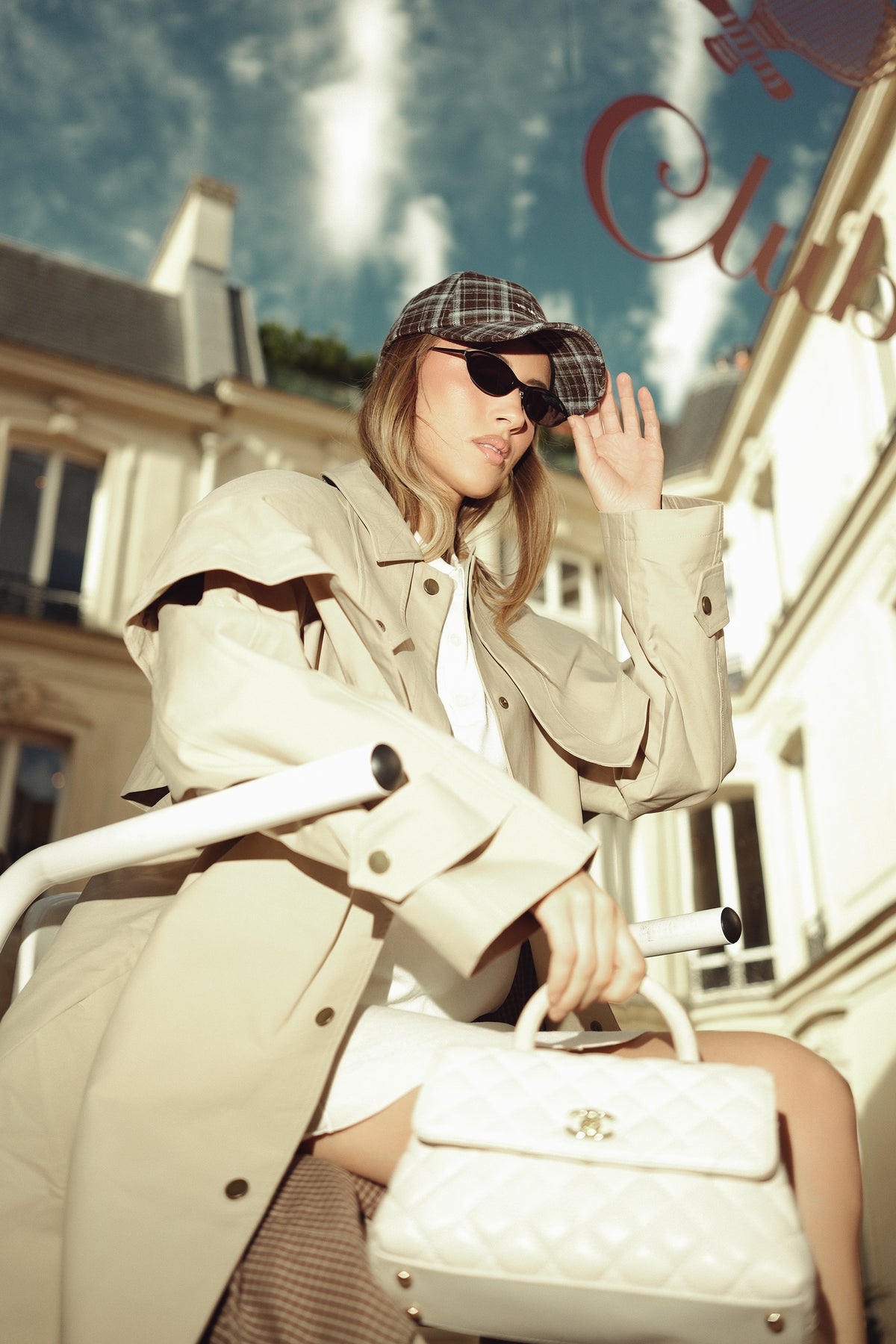 Model wearing TUK Chestnut Blue Check Heritage Plaid Cap with black sunglasses, beige trench coat, and chic white quilted handbag.