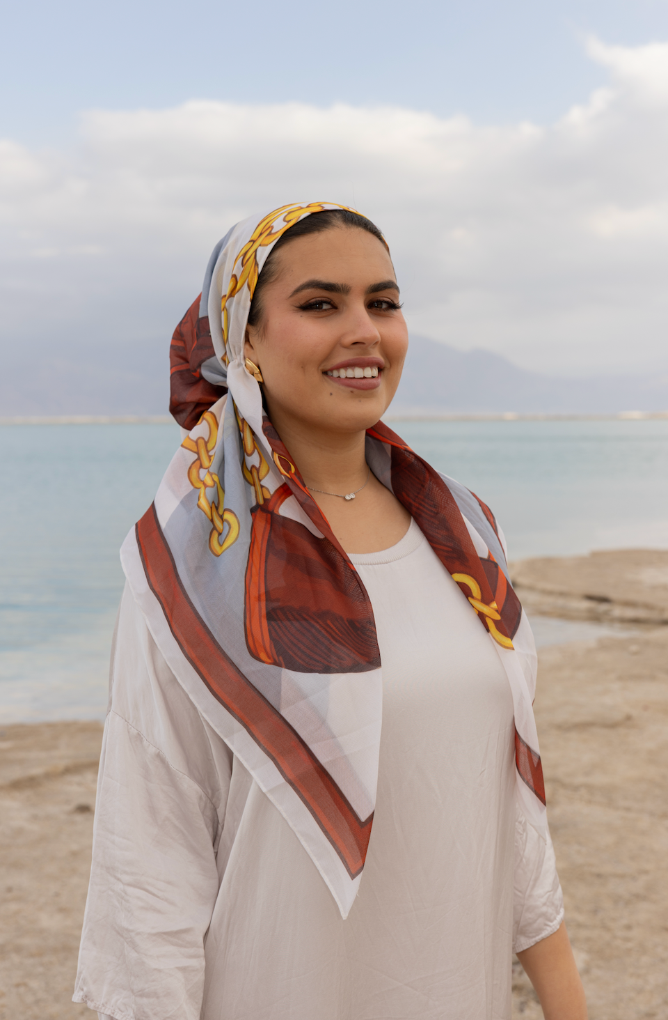 Woman wearing TUK Cool Reins head scarf by the sea, styled in elegant patterned silk with neutral outfit and soft natural backdrop.