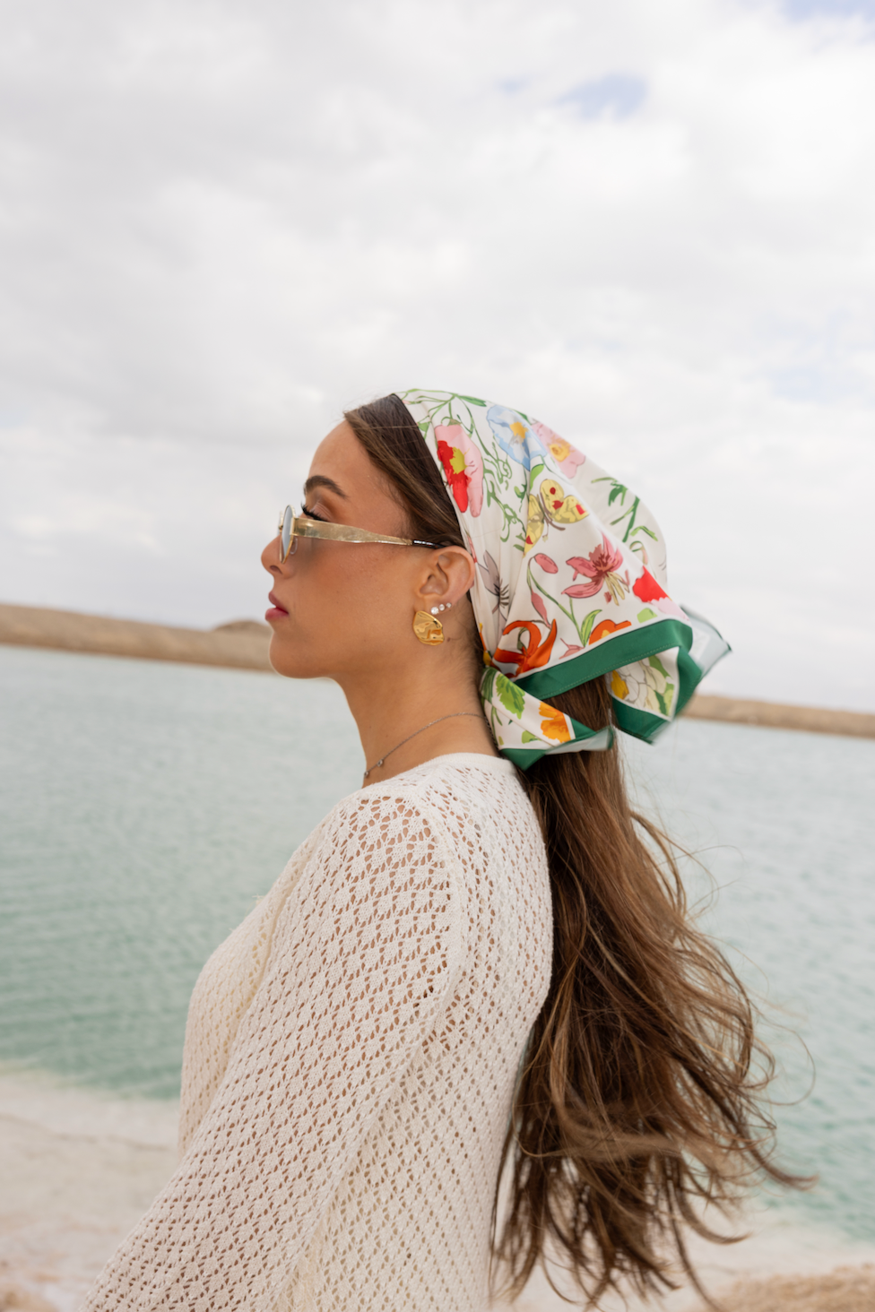 Woman by the sea wearing TUK Field Day Silk Scarf, a colorful floral headscarf styled with sunglasses and coastal elegance.