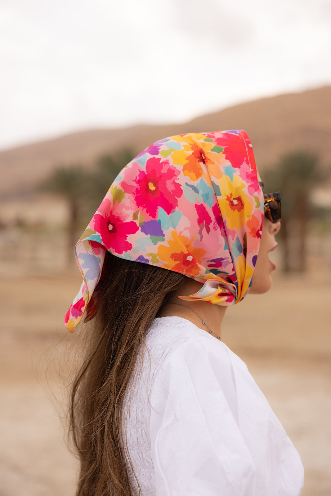 TUK Flower Hour Silk Bandana with vibrant floral print styled as a silk headscarf, paired with sunglasses in a desert setting.