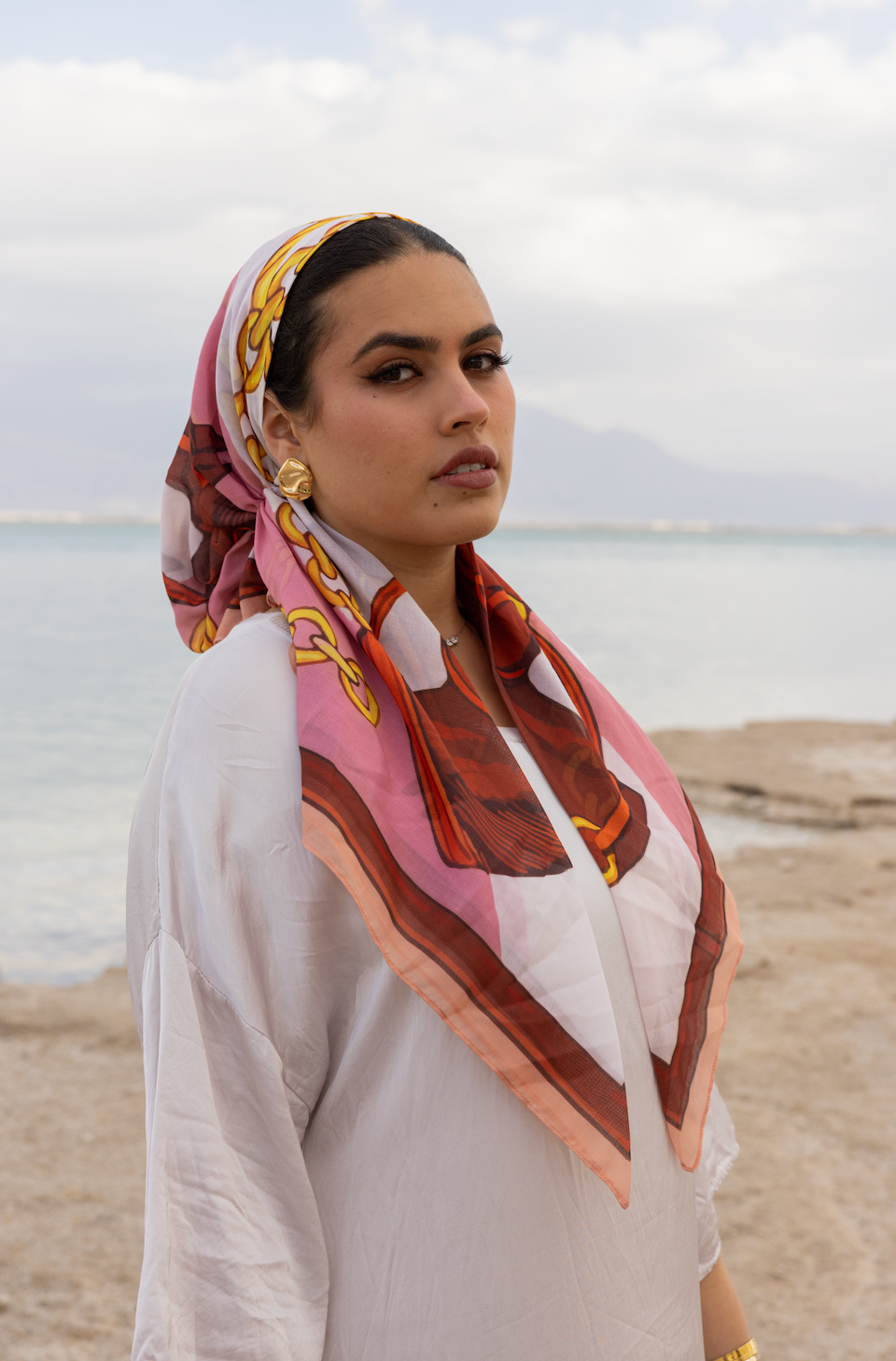 Elegant woman by the sea wearing TUK Giddy Up Gorgeous Head Scarf, a vibrant silk accessory with bold equestrian-inspired design.