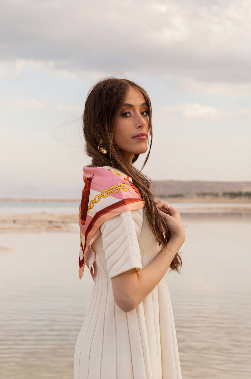 Model wearing TUK Giddy Up Gorgeous Silk Scarf, pink equestrian print, styled over dress by serene beach at sunset.
