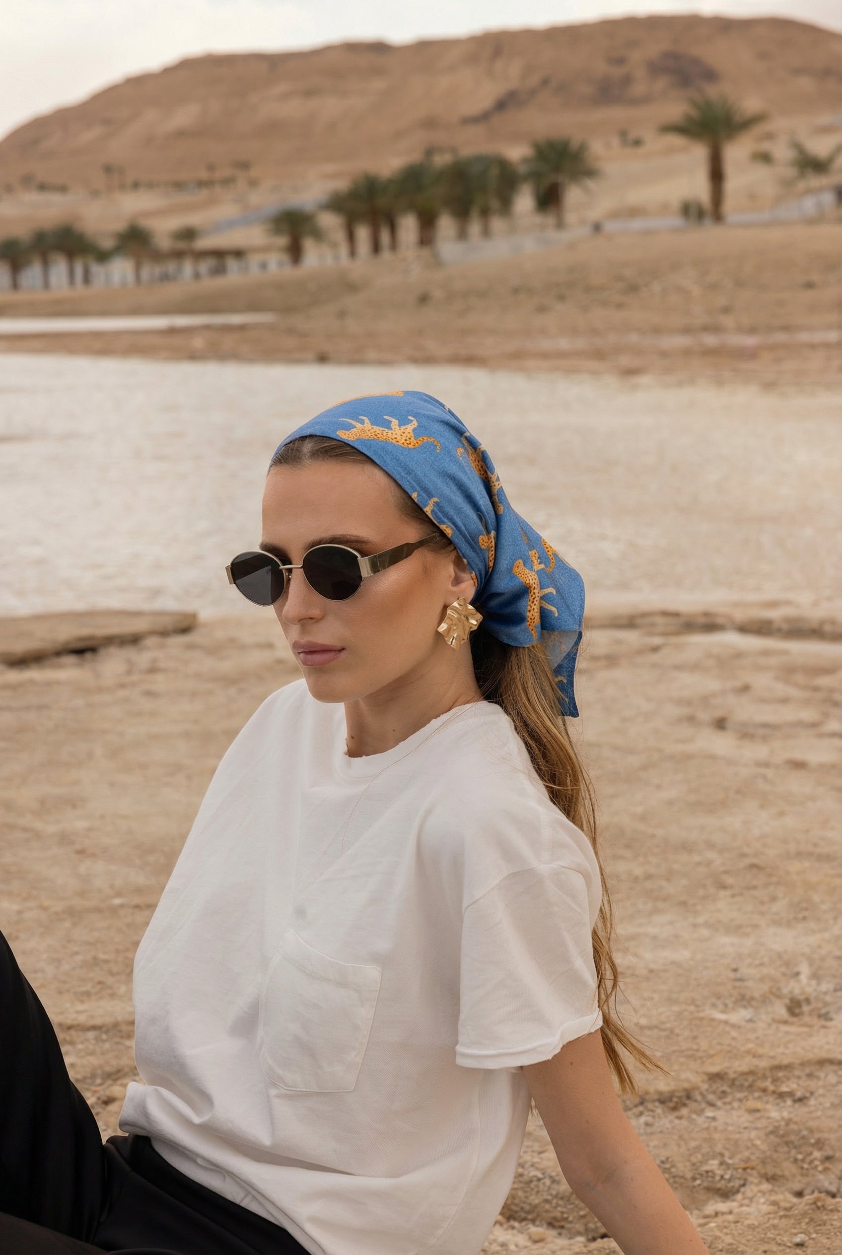 Model wearing TUK Haute Pursuit Silk Scarf crochet bandana in blue leopard print at a chic desert beach setting.