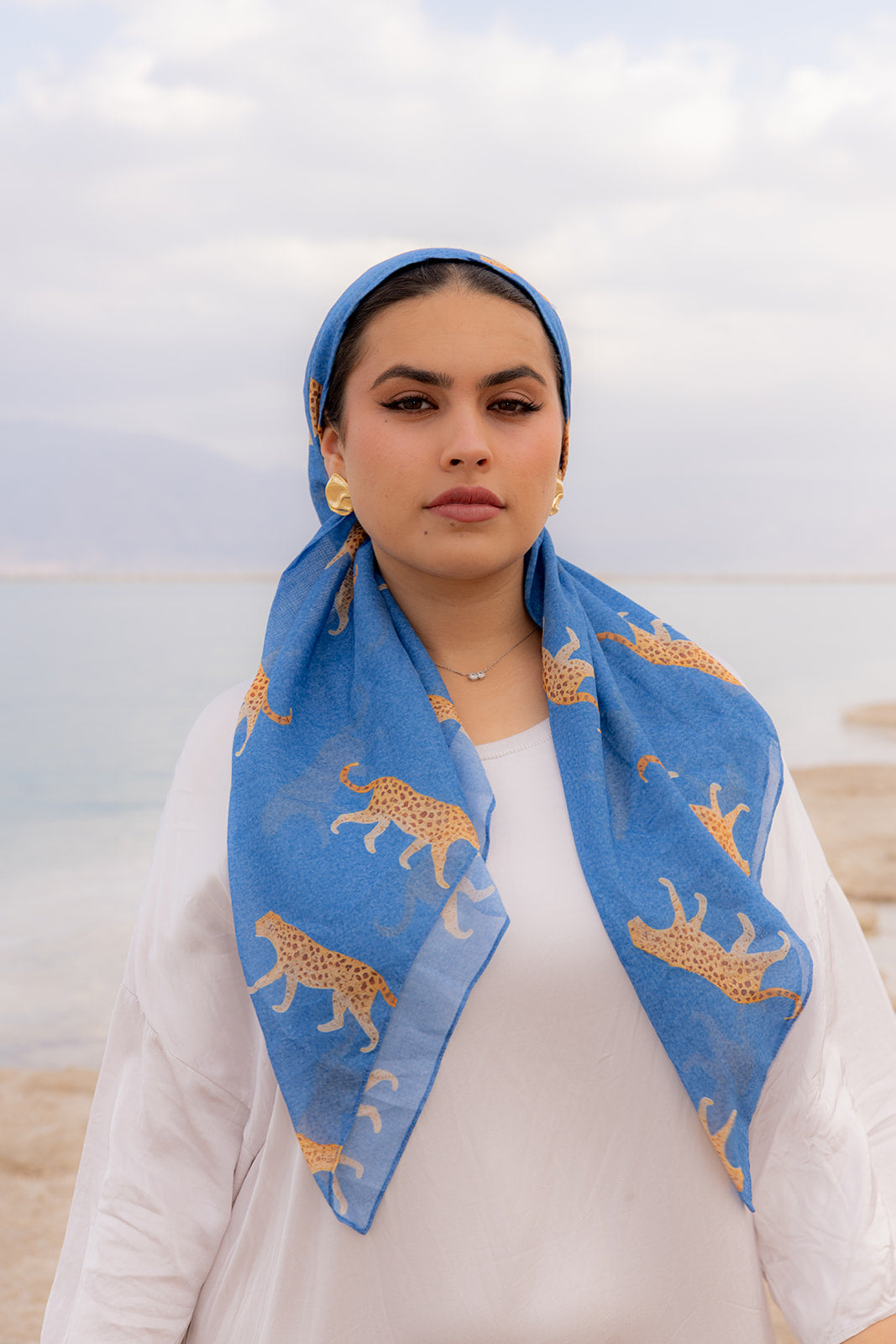 Haute Pursuit Head Scarf by TUK in vibrant blue with golden leopard print, elegantly styled by a woman by the water.
