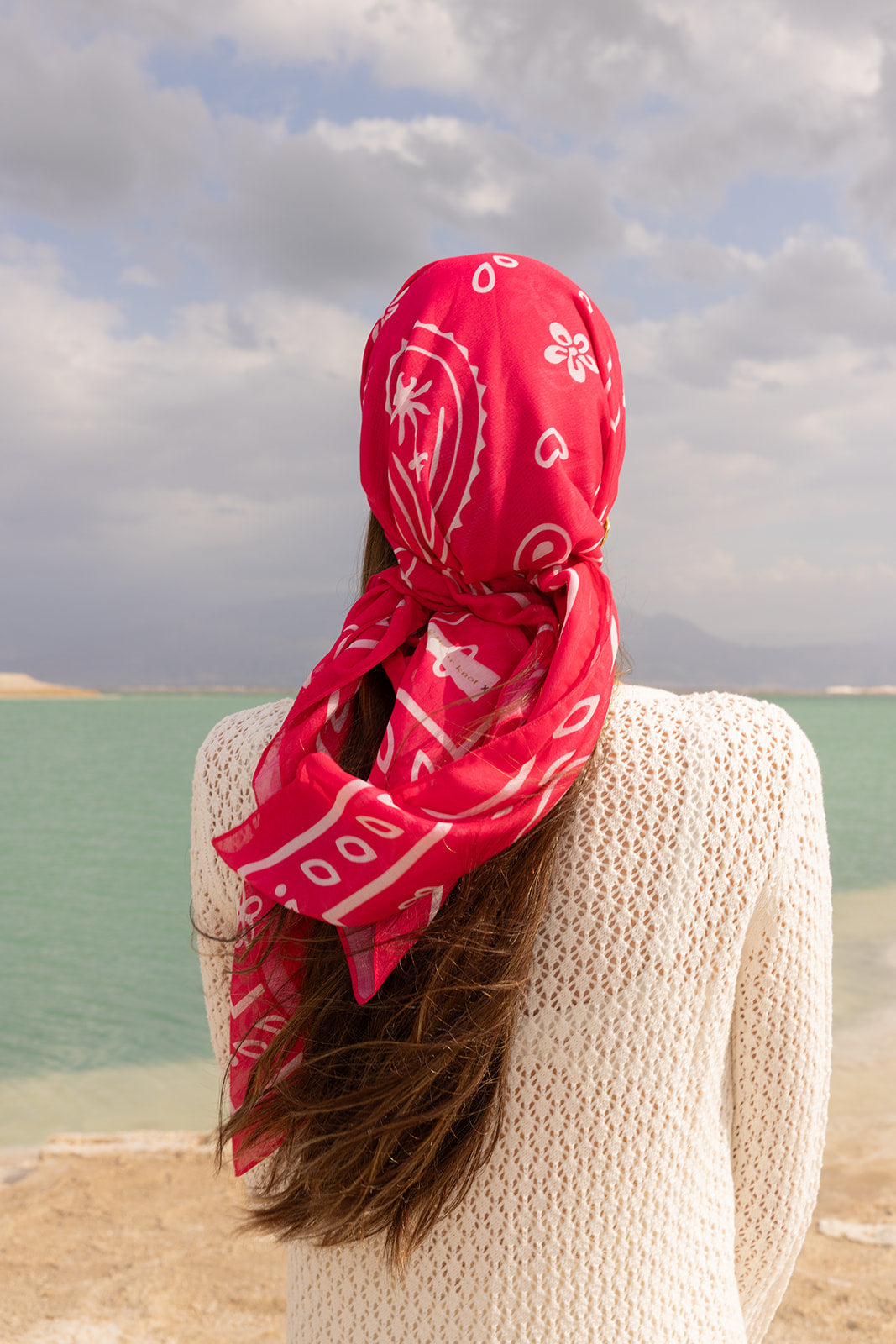 Knot Today Head Scarf by TUK in vibrant pink paisley print, styled as a chic head wrap by a serene turquoise shoreline.