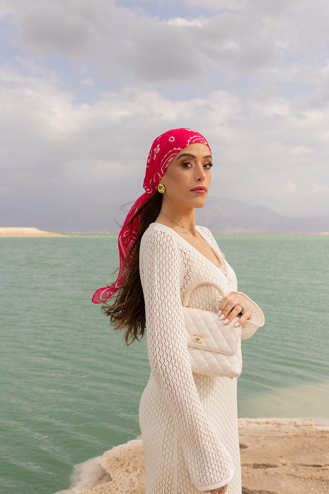 TUK Knot Today Head Scarf in vibrant pink paisley print, styled as a chic head wrap beside a tranquil turquoise coast.