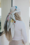 Woman with long wavy blonde hair wearing a white silk headscarf featuring a watercolor butterfly and botanical print, tied at the back with long flowing ends, standing beside a sunlit wall.