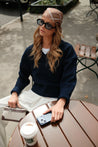 Model wearing TUK’s Neutral Position headscarf with navy sweater and white pants, sitting at café table with coffee.