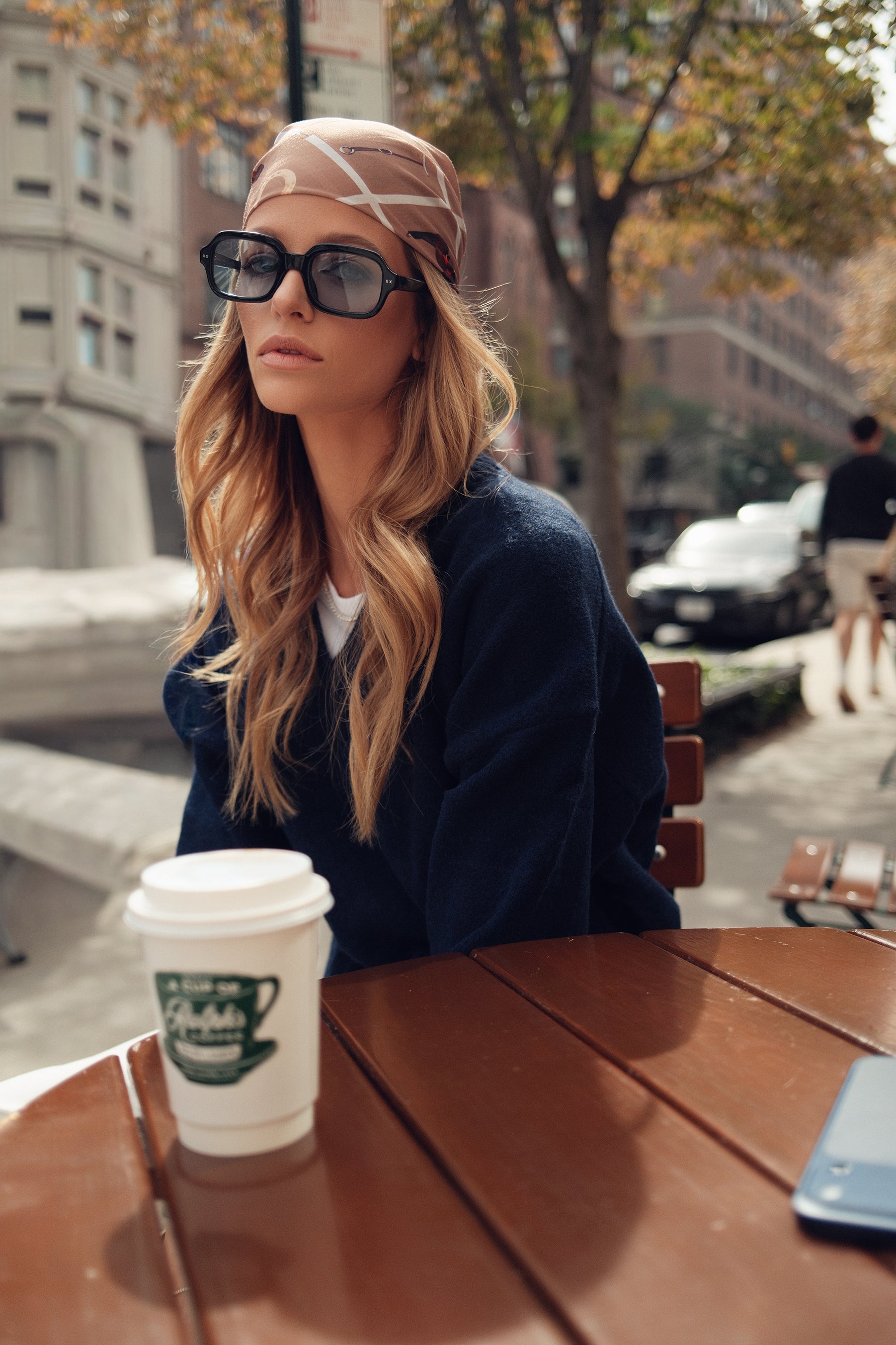 Woman sitting at an outdoor cafe table with a coffee cup, wearing sunglasses and a headscarf.
