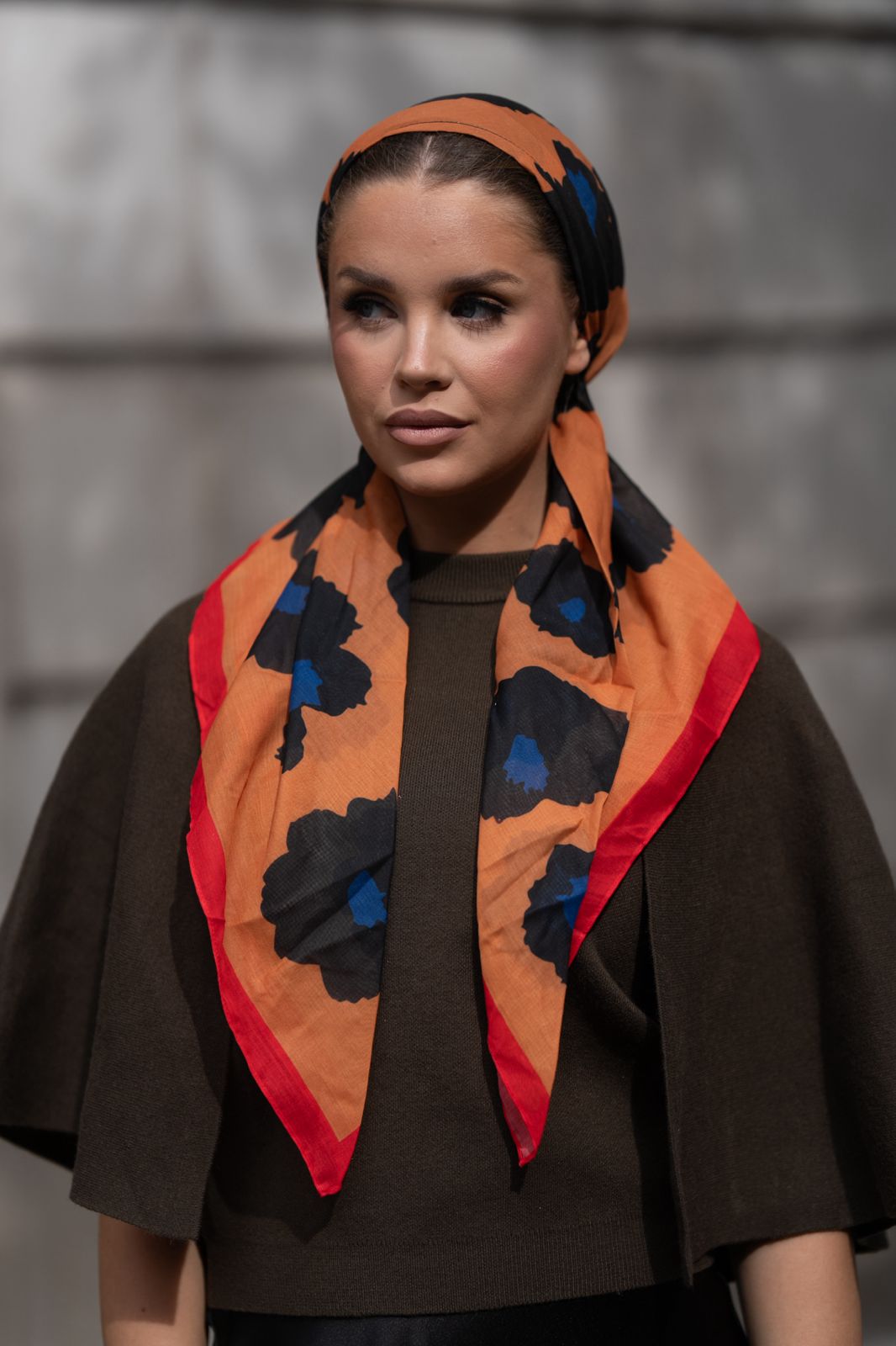 Model wearing TUK’s Out Of Line Pre-tied Head Scarf in bold orange with black and blue floral patterns and red border.