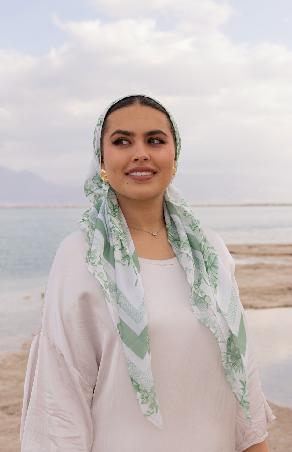 Woman wearing TUK Sage Advice headscarf in green and white, elegant modest fashion accessory by sea under cloudy sky.