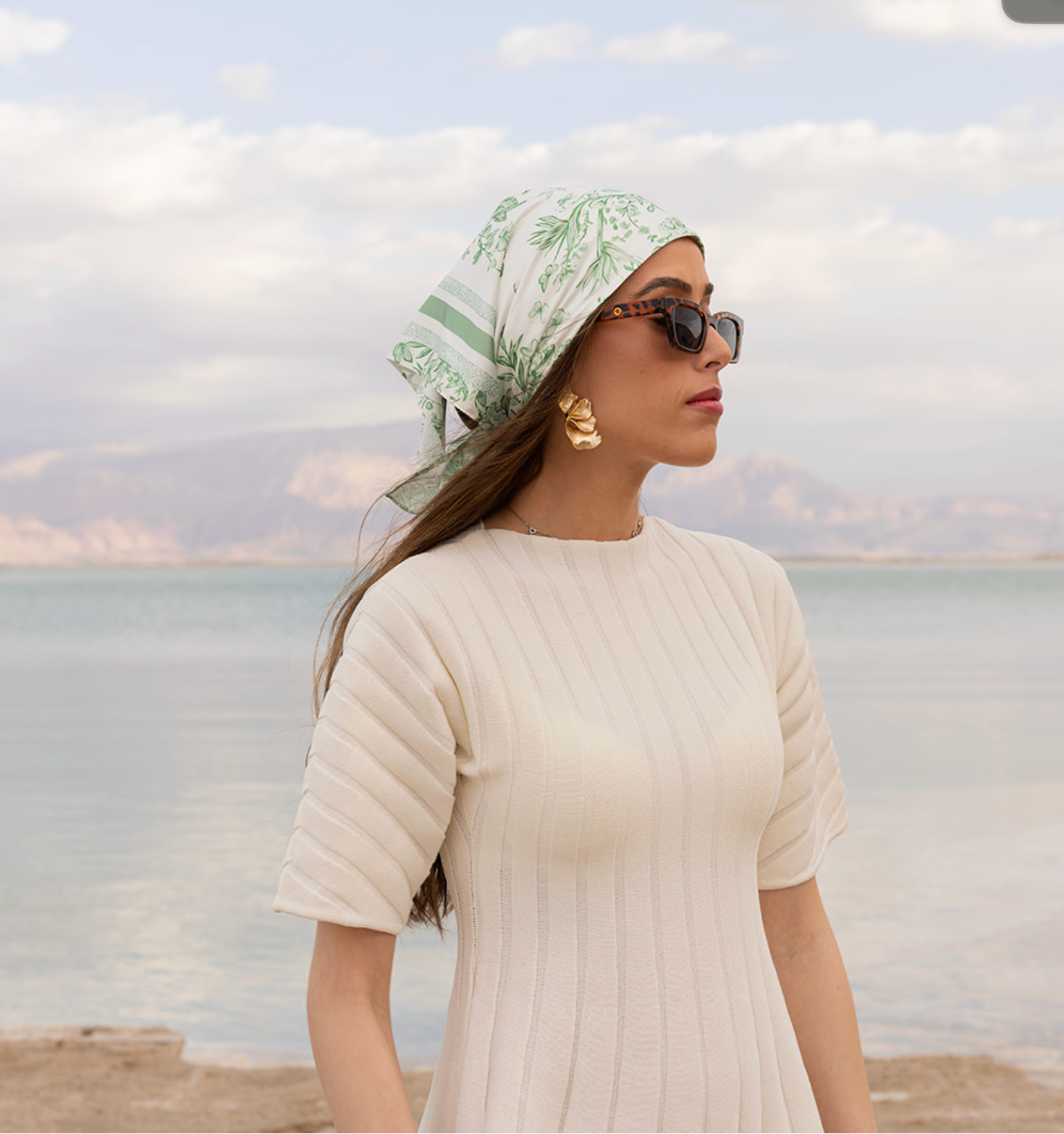 Woman by the sea wearing TUK Sage Advice Silk Scarf, green botanical print, styled with sunglasses and neutral dress.