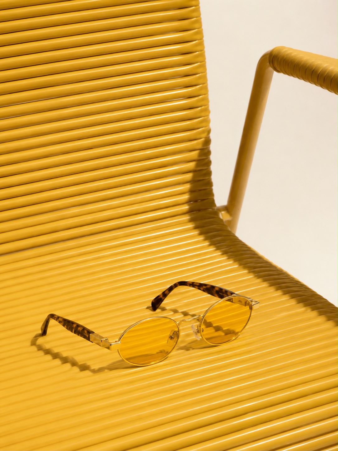 Sola Sunglasses by TUK featuring oval yellow-tinted lenses, gold metal frames, and tortoise temples on a modern yellow chair.