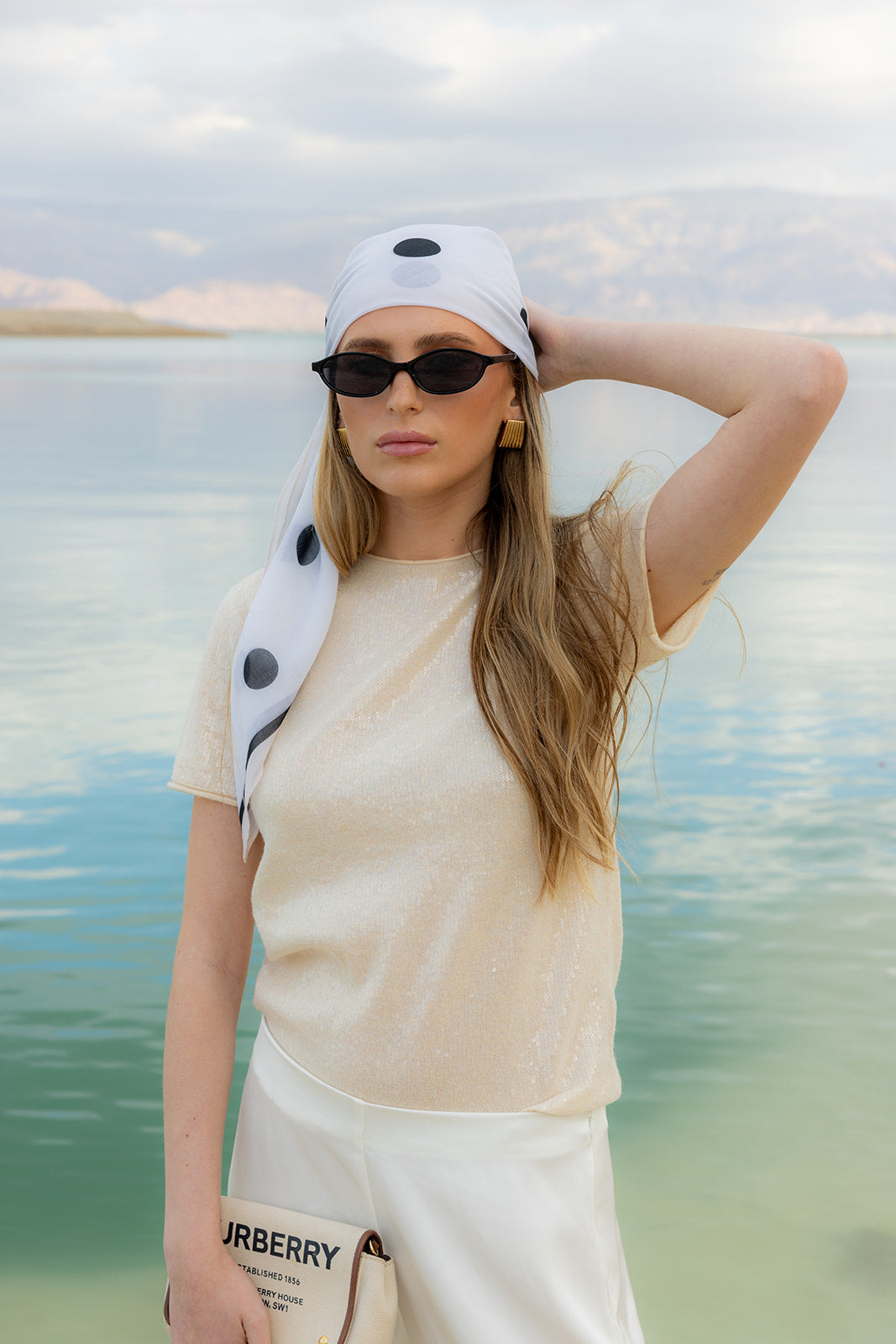 Model wearing TUK Spot On Silk Bandana in white with bold black polka dots, styled as a chic headscarf by a serene lakeside.