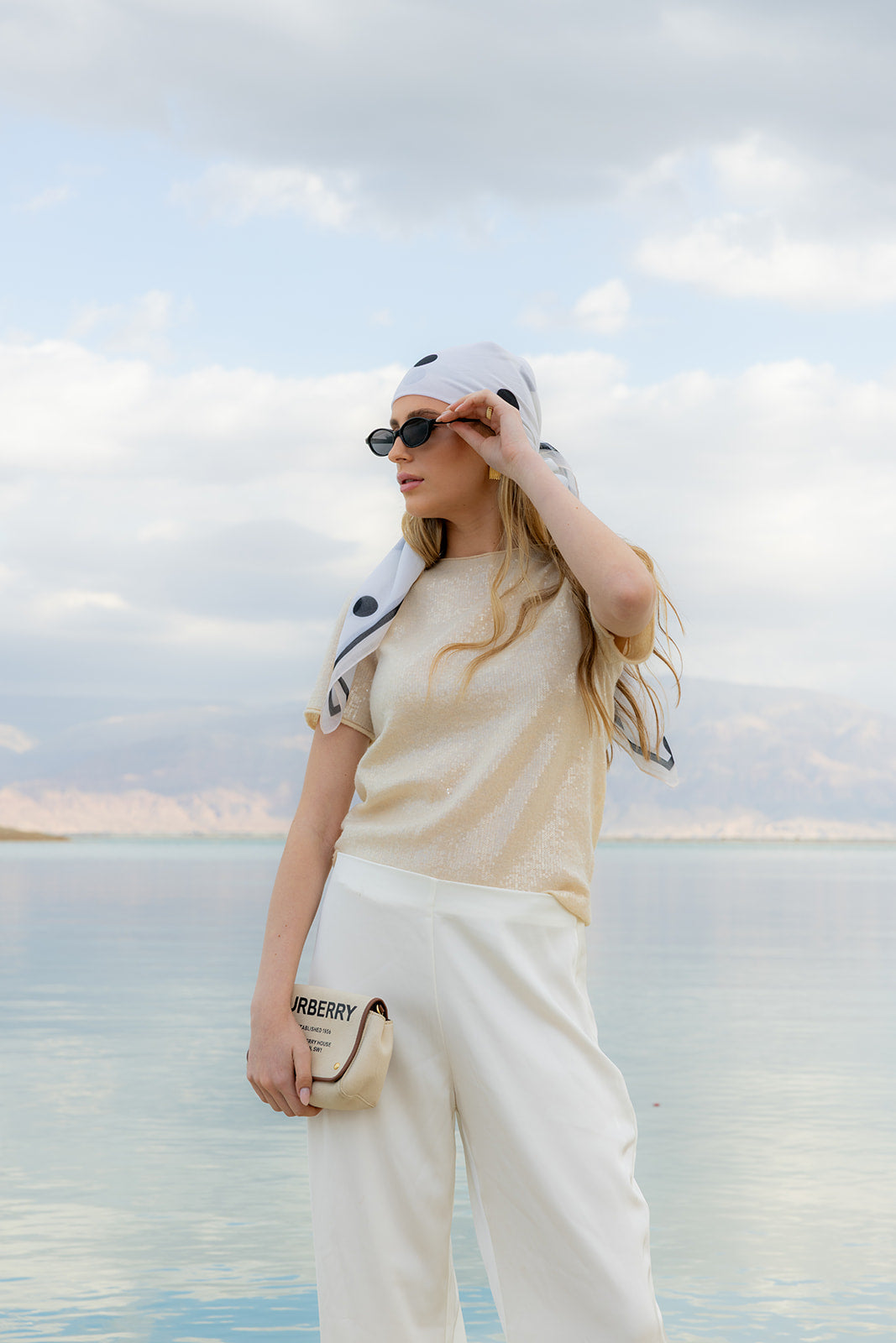 Model wearing TUK Spot On Silk Bandana as a stylish headscarf in white with black polka dots by a tranquil lakeside view.
