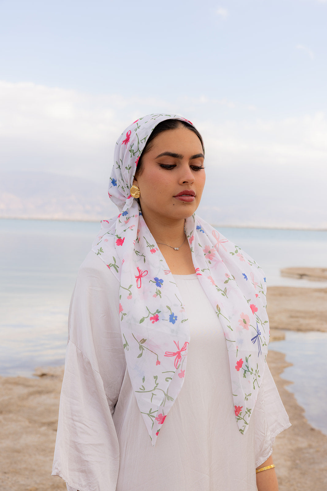 TUK Spring Thing Head Scarf crochet bandana in a white floral print, styled by a woman in a peaceful coastal setting.