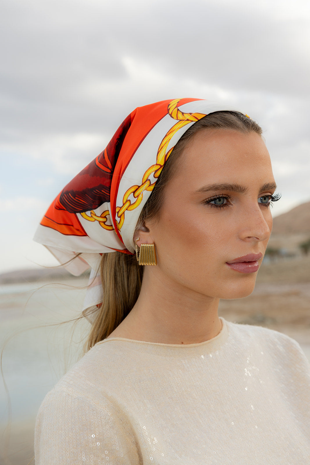 TUK Stirrup Society Silk Bandana with bold equestrian chain print styled as a chic headscarf by the seaside in cream attire.