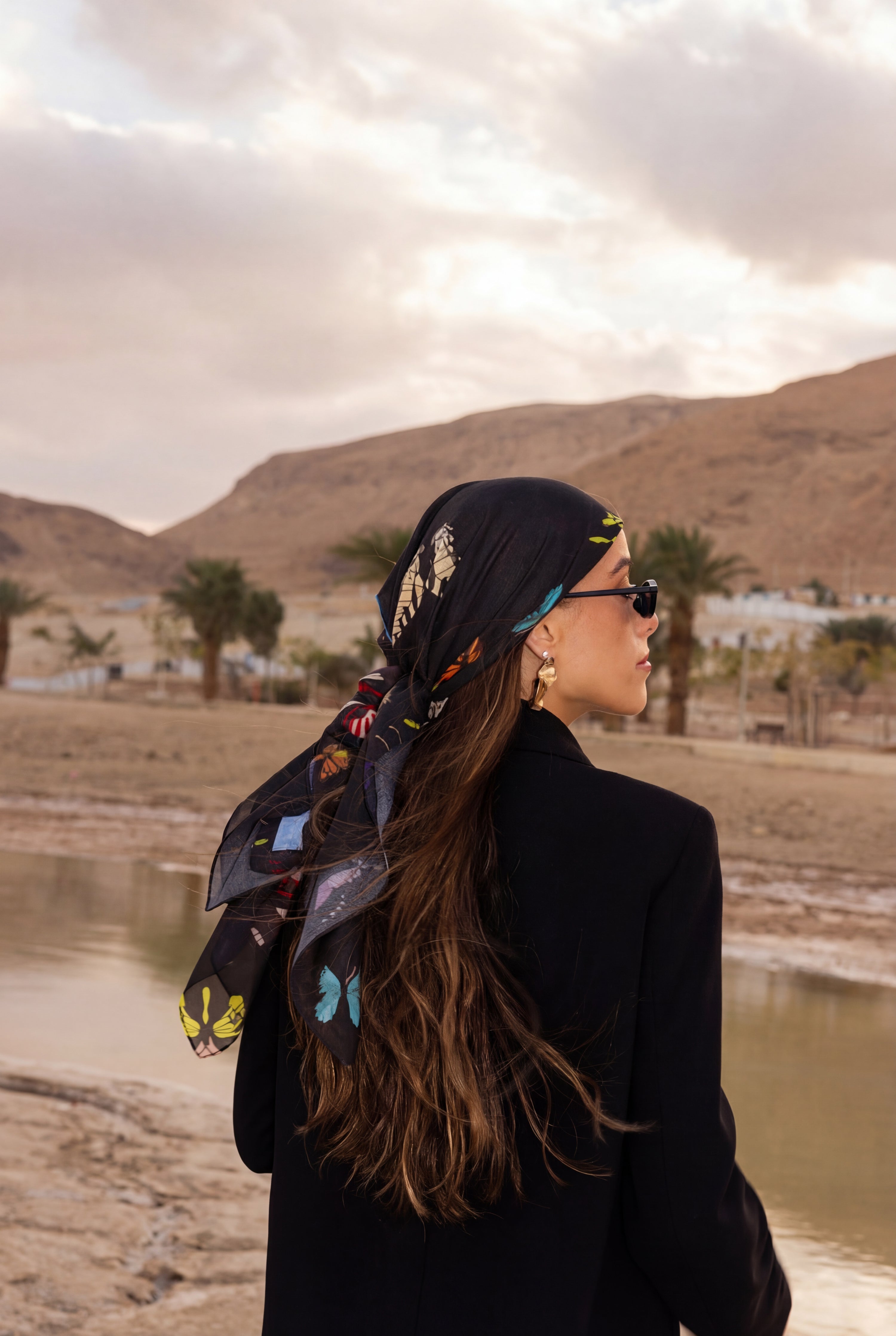 Woman wearing TUK The Night Shift headscarf, black with colorful butterfly print, styled in desert landscape with mountains.
