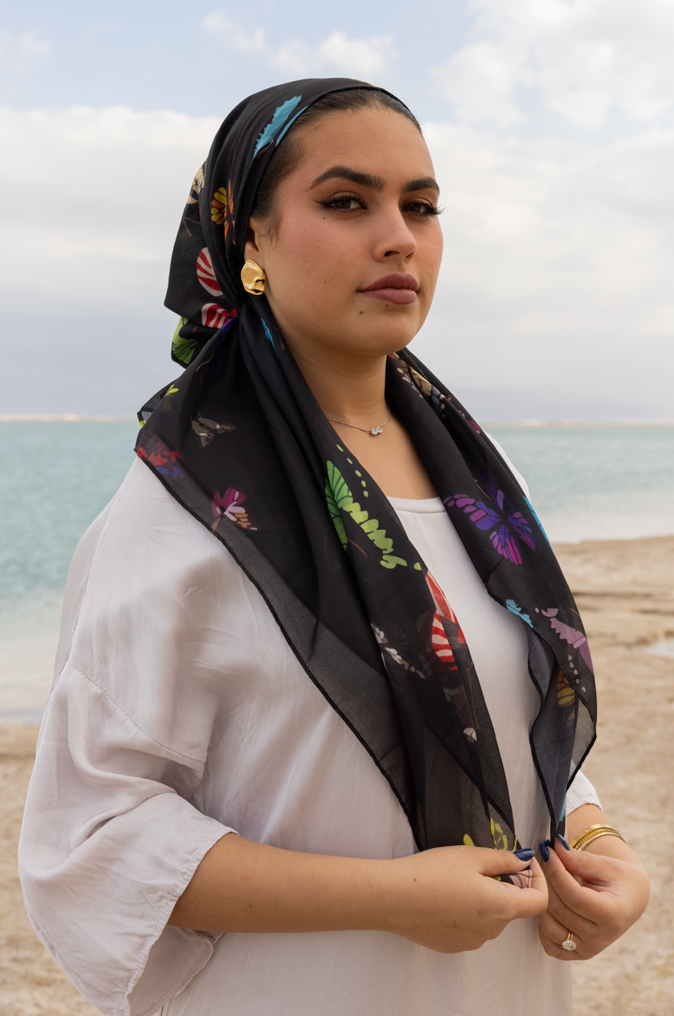 Woman wearing TUK The Night Shift headscarf, black butterfly print scarf styled by the sea, elegant modest fashion look.