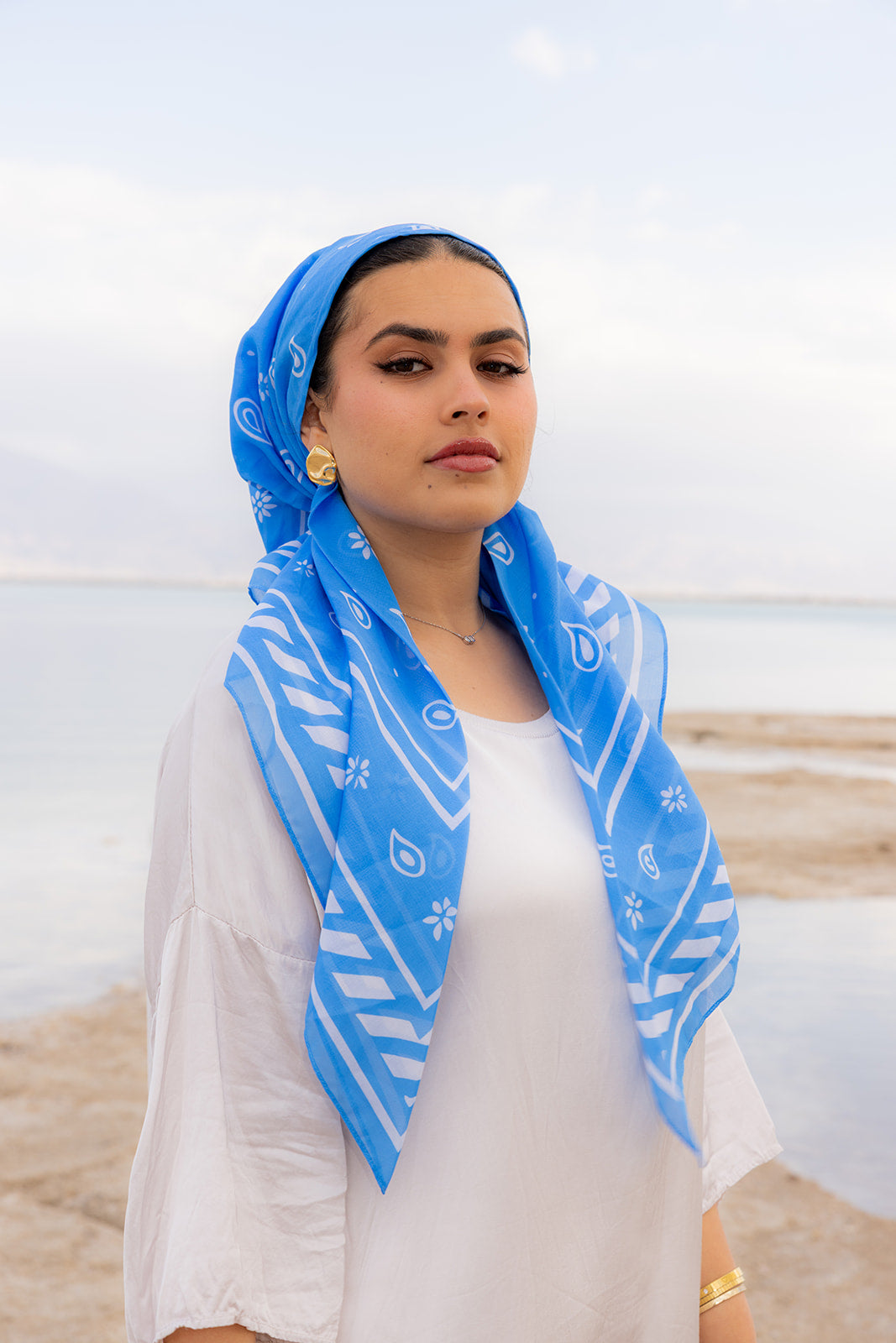 TUK Tied & True Head Scarf crochet bandana in sky blue paisley print, styled by a woman in a serene coastal setting.