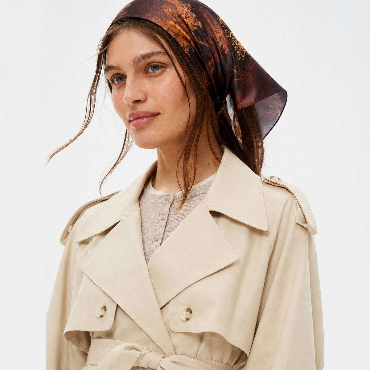 Branch Out Silk Bandana on a person in a trench coat, showing the dark brown, orange, and gold nature print.